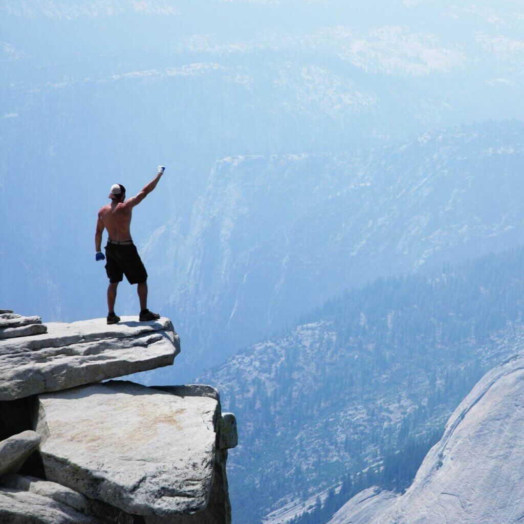 A man standing on a cliff edge, waving at a breathtaking view of the mountains.
